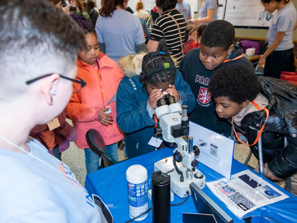 A group of young Black children take turns looking into a microscope. 
