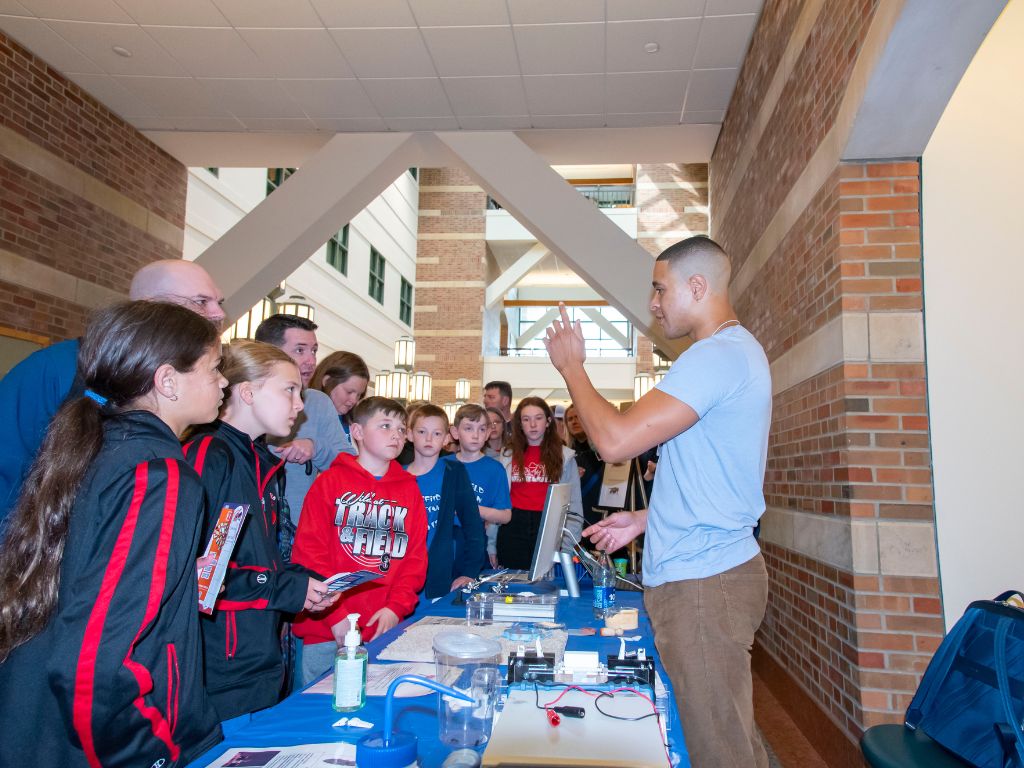Students standing at a table staring at a man in a blue t-shirt and khaki pants. 