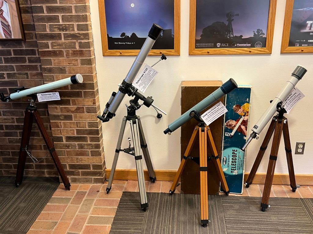 A line of telescopes are set up against a brick and painted white wall. There are framed pictures of a dark sky behind them.
