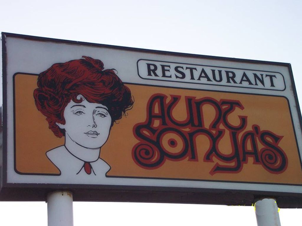 A large sign for Aunt Sonya's restaurant. The words Aunt Sonya's are written in dark blue curvy letters and orange outlines and on the left is a portrait of a white woman wearing a collar and her orange curly hair piled up on her head.