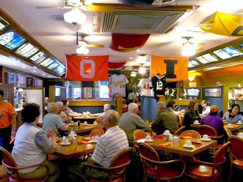 the inside of aunt Sonya's restaurant. Ohio State and University of Illinois flags hang from the ceiling. People are seated at tables in the middle of the space and in booths against the left wall. On the right is a counter where you can see chefs making food and handing it to servers. 