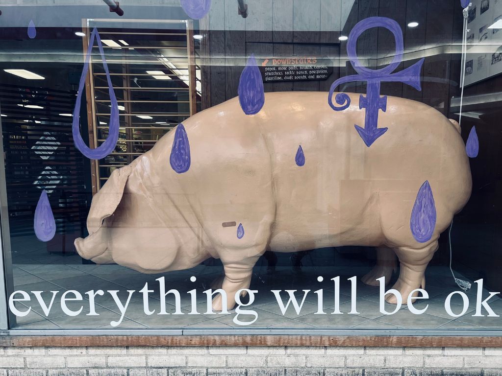 A glass window display that says everything will be ok with purple raindrops and a life-size pink pig standing in the center.