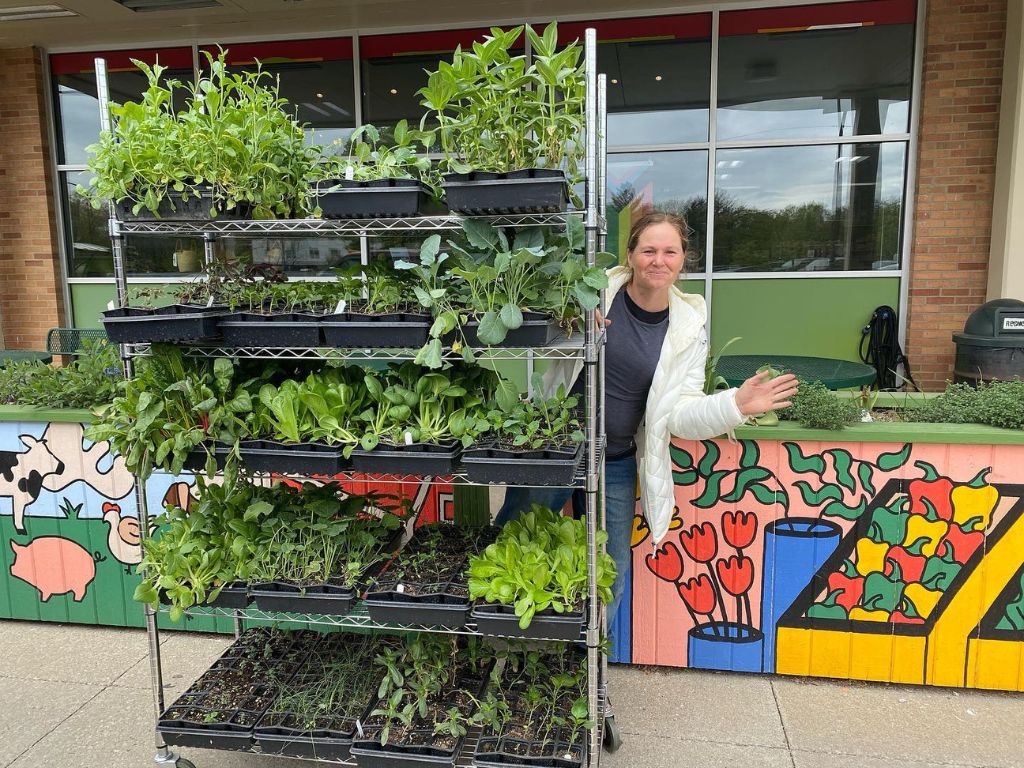 A white woman in a blue t-shirt and white zip up hoodie waves from behind a tall metal shelf full of plants. The shelf is in front of a a wall of windows and a low wall that is painted with colorful flowers and veggies.