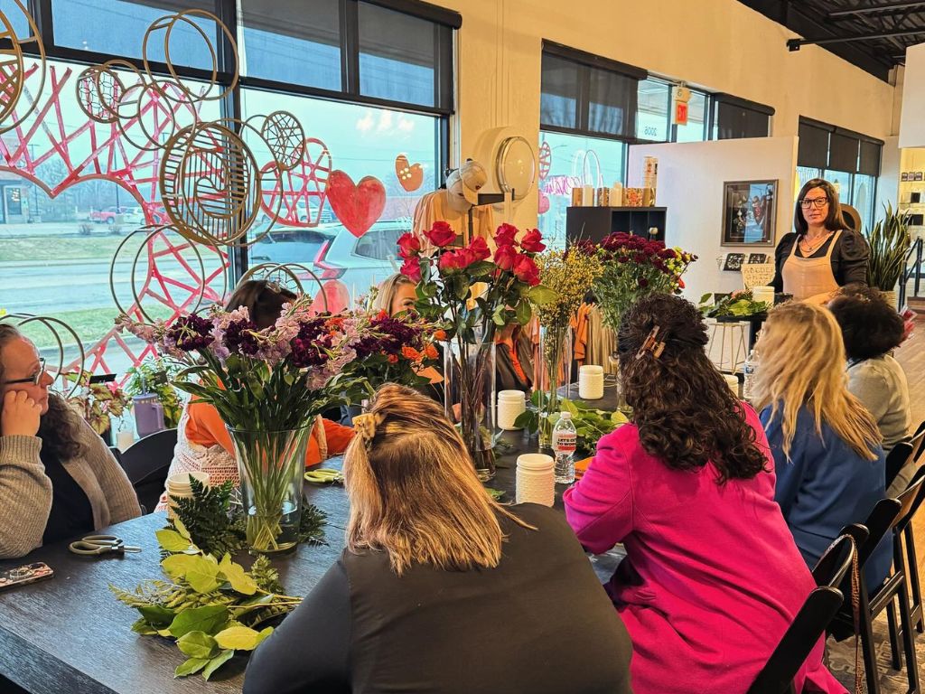 A group of people sit around a large table with flowers on it in front of a window watching an instructor in the front of the group
