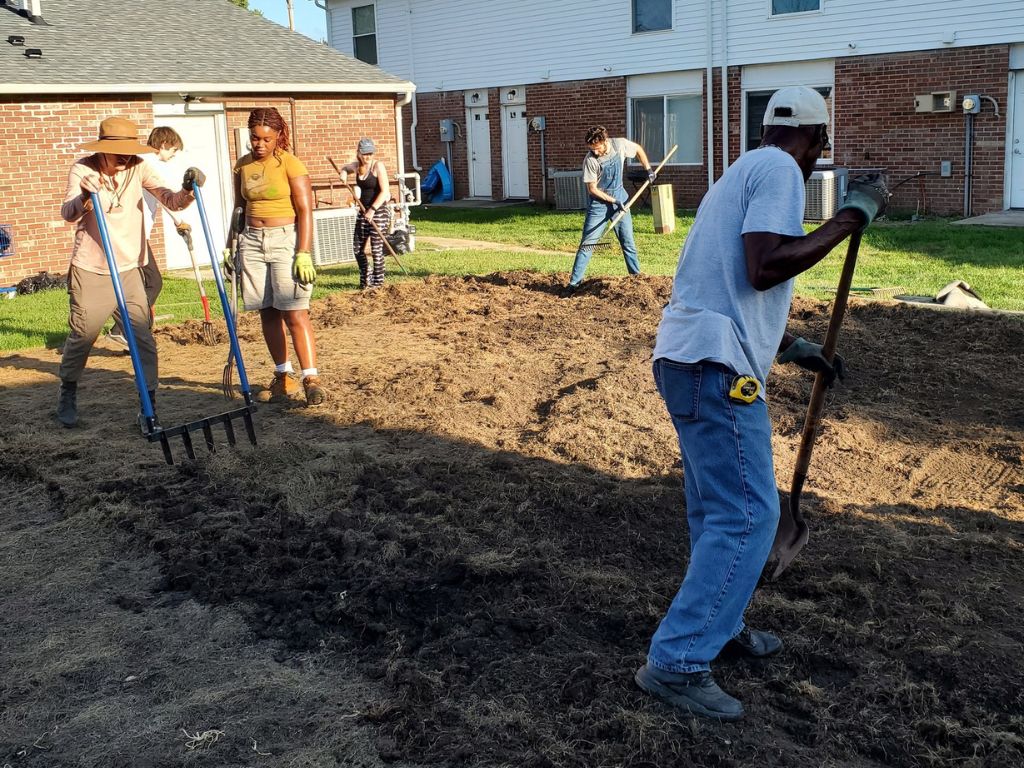 People stand on an empty plot of dirt transforming it into usable soil. you can see the back of a black man in a white t-shirt and jeans holding a shovel to dig on the right side. on the left are 5 other people using various tools to dig up the soil. In the background are two level apartment buildings. 