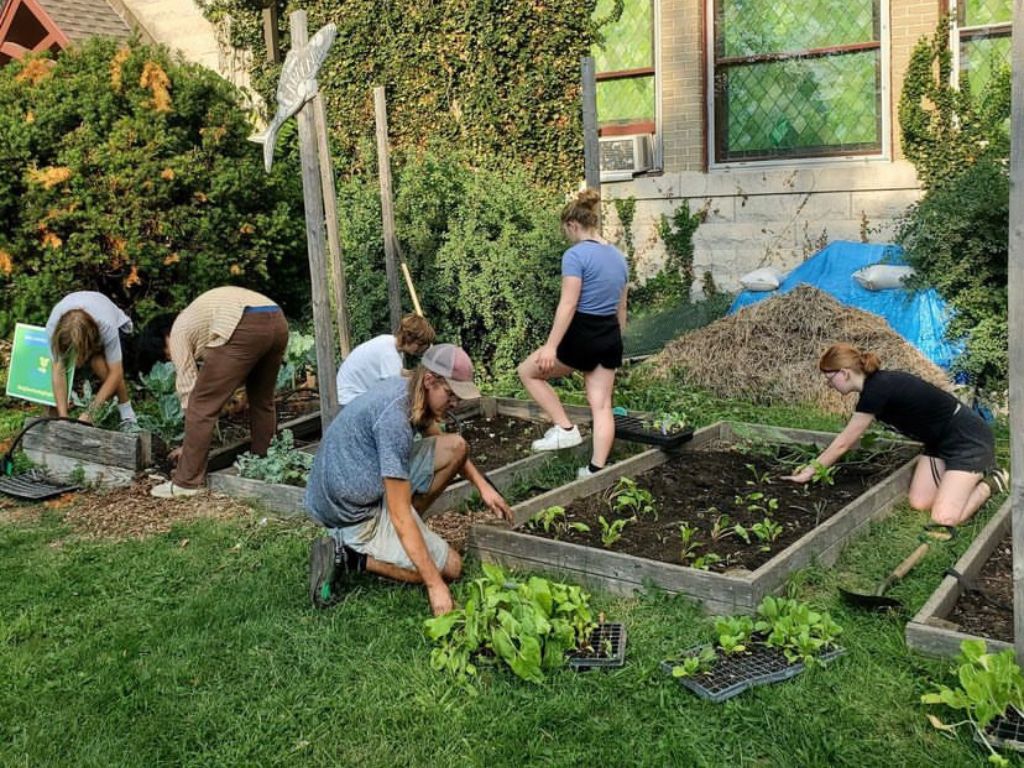 A group of people work in two small plots of land near a gray brick building. 
