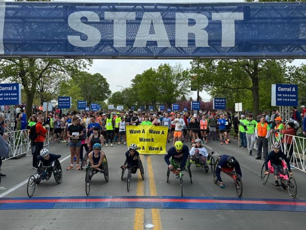 Under a giant blue banner with the word START in capital letters. Beneath are seven athletes in wheelchairs in the start position. Behind them are thousands of racers.