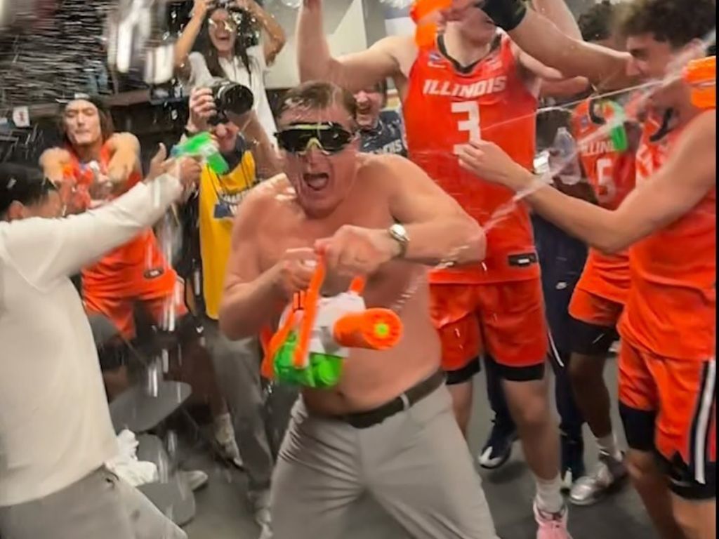 A shirtless white man in goggles and gray shorts sprays a water cannon at the camera in a locker room celebration. He is surrounded by players in orange jersey's shooting water guns and throwing water bottles.