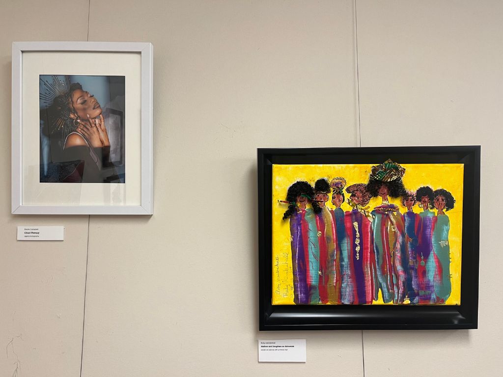 A tan wall with two art pieces framed on the wall. One the left is a portrait of a black woman wearing a golden crown with her hands on her face. On the right is a mixed media painting with a yellow background and figures standing together in colorful clothing with real hair attached to the canvas.