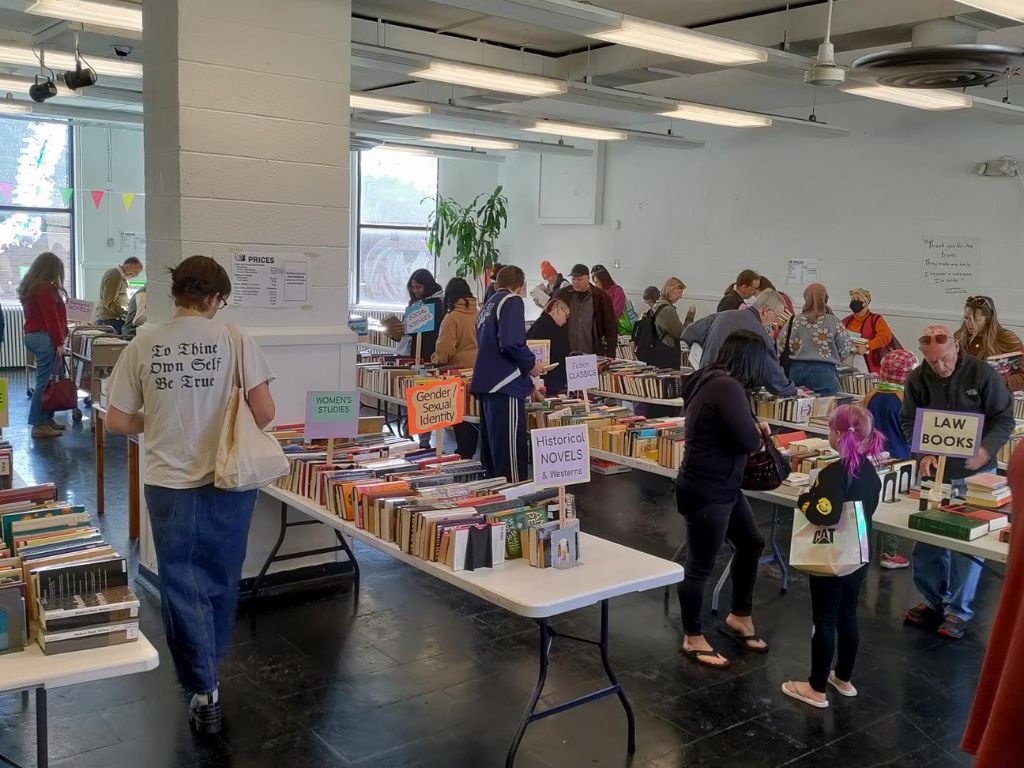 An open space full of tables of books and people walking along the tables. There are bright florescent lights above and a window on the left wall.