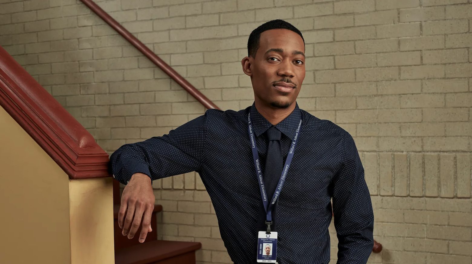 A Black man wearing a collared shirt and a lanyard stands with his arm against a stairs railing looking at the camera