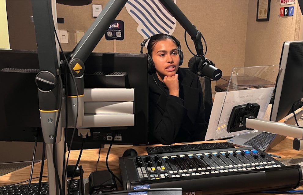 A Black woman sitting behind a microphone reading a prompt for the radio