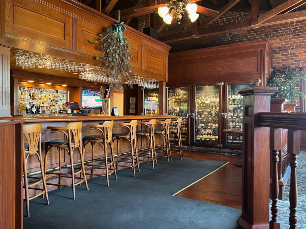An empty bar with a peacock feather decor above the barstools. A wooden cabinet with full-size glass doors holds a variety of wine bottles.