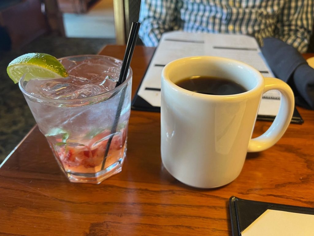 A glass cup with a lime wedge and two black straws with muddled strawberries at the bottom beside a white mug of black coffee.