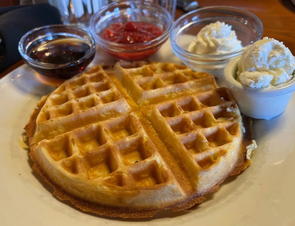 A waffle with side cups of syrup, strawberries, whipped cream, and butter.