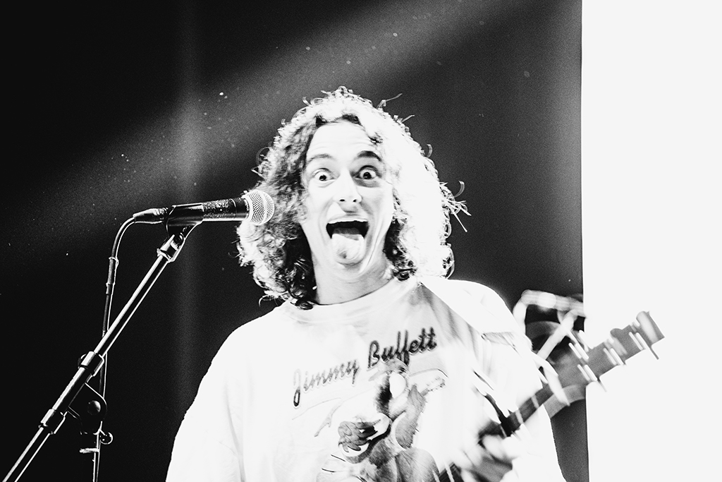A guitarist with long, curly hair is making a lively facial expression while playing an electric guitar. The musician is wearing a t-shirt and appears to be in a moment of joyful performance, contributing to the lively atmosphere of the concert.