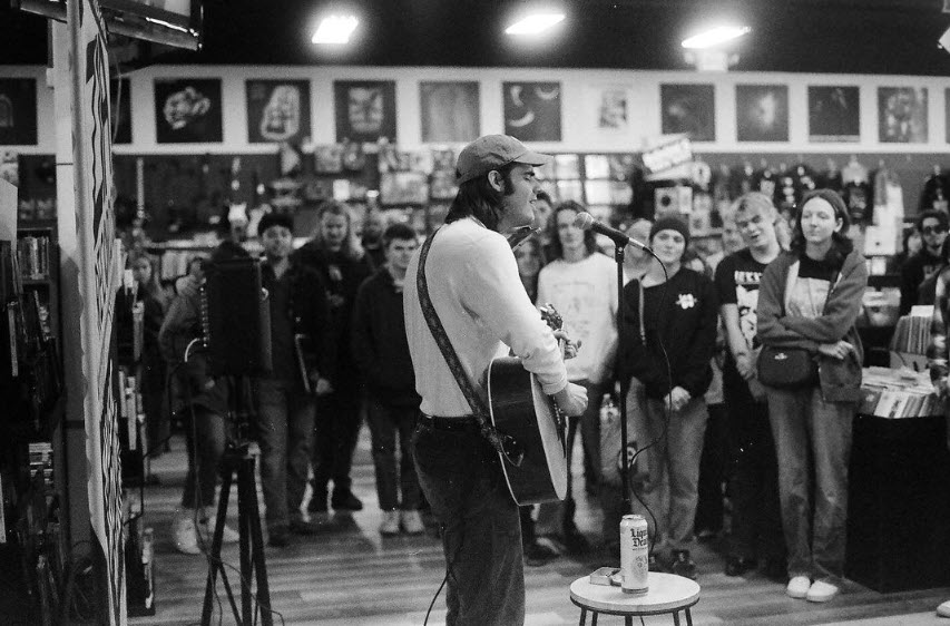 A black and white photo captures a musician performing in a record store. The artist, wearing a cap and a casual outfit with a guitar strapped on, sings into the microphone, with focus and emotion evident in his posture. The audience, an attentive and diverse group of individuals, some with discernible smiles, stand among the racks of records, their attention drawn to the performer. The image, with its grainy texture and candid feel, offers a glimpse into an intimate live music scene, resonating with the charm of a bygone era.