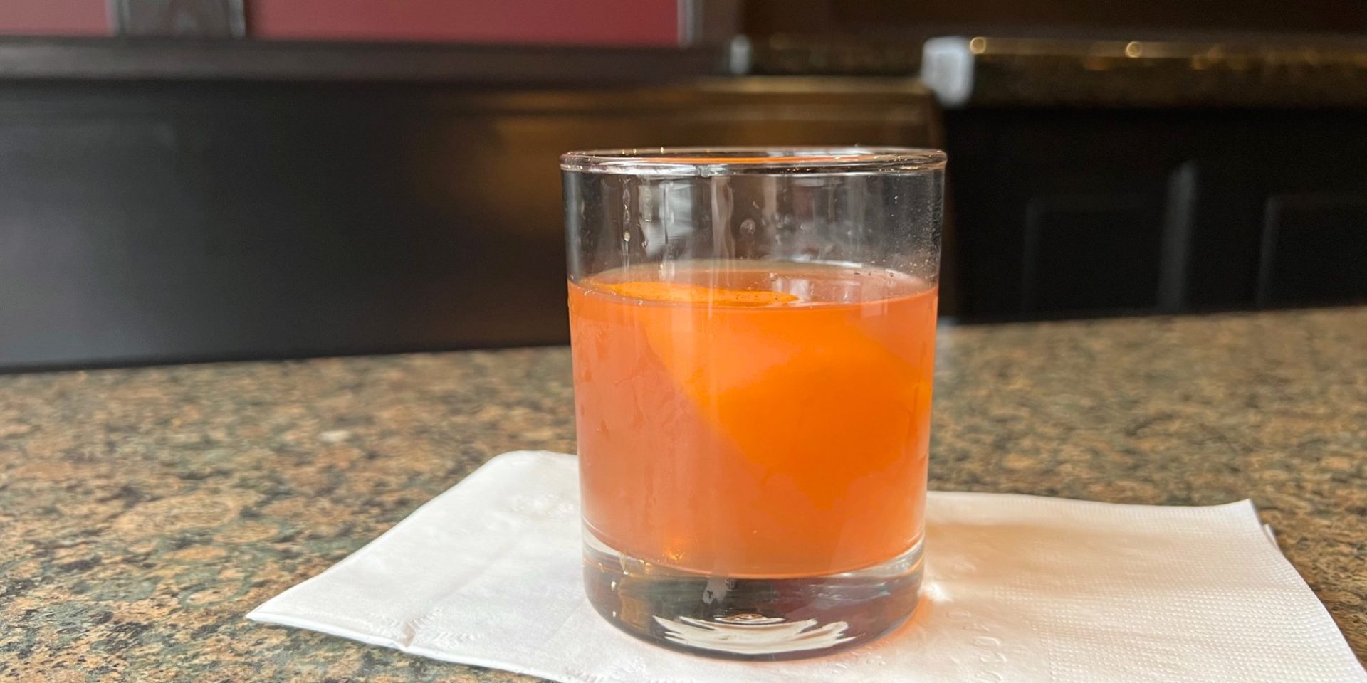 A redish pink cocktail with an orange peel in a half rocks glass on a white napkin on a granite tabletop during the daytime.