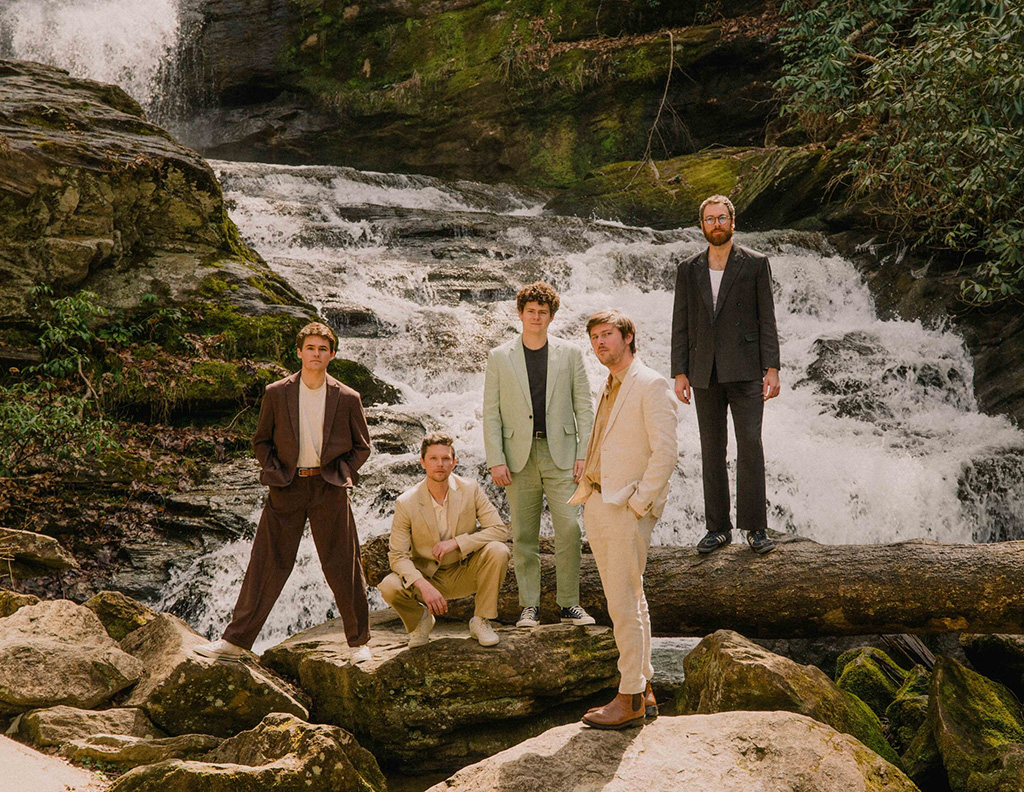 In a scene staged at a cascading waterfall, the band, The Brook and the Bluff, are captured in a moment of repose against nature's grandeur. The members are smartly dressed in a relaxed, yet stylish assortment of suits and casual attire, evoking a sense of laid-back sophistication.