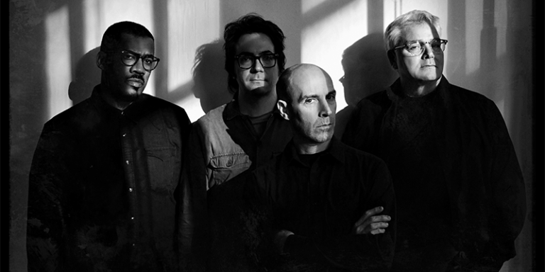 four men positioned against a backdrop with shadow patterns created by light filtering through a window or similar object. From left to right: the first man wears glasses and a dark shirt, adopting a serious expression. Next to him, a younger man with shoulder-length hair and glasses is dressed in a casual button-down shirt, also looking intently at the camera. The third man, bald and with a distinct focused gaze, is clad in a dark, simple top. The final figure, an older man wearing glasses, is slightly blurred compared to the others, suggesting some movement or a focus shift in the image, and he is also dressed in dark colors. They all wear dark, muted clothing, giving the group a cohesive, solemn appearance.