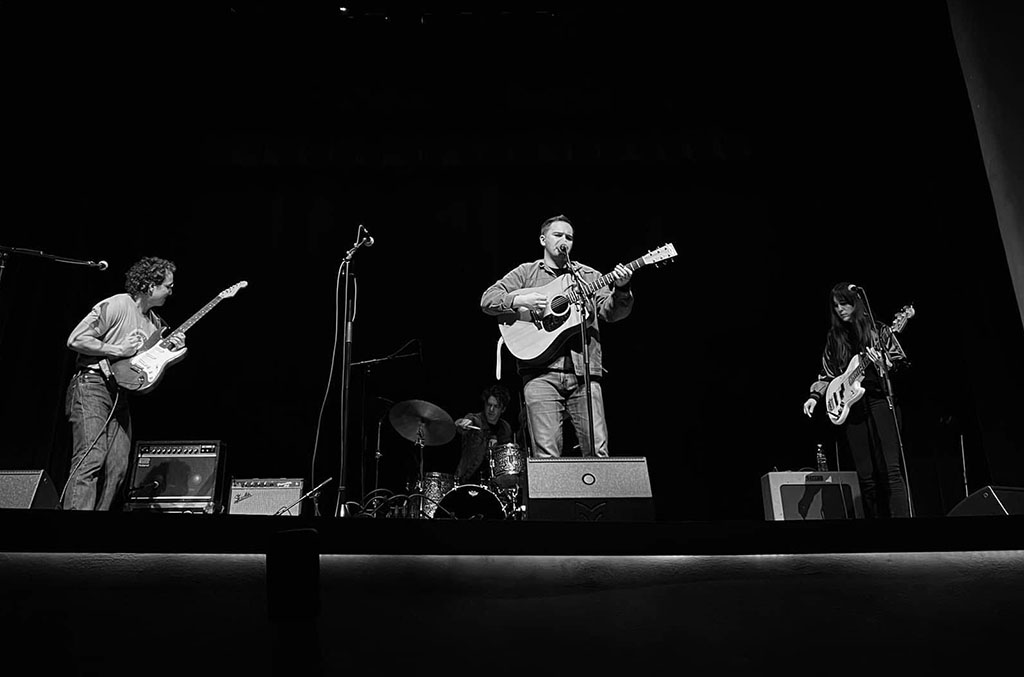 A black and white photo featuring 3 band members onstage performing.