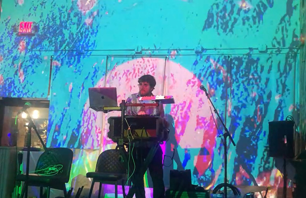 The artist is seen from a performing in a dimly lit room with vibrant, psychedelic projections on the wall behind. The light casts a colorful glow across the space, enhancing the immersive experience of the performance.