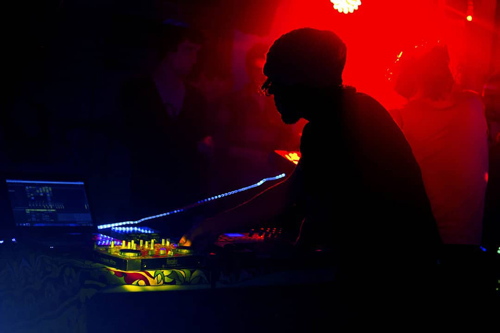 An intimate club setting, with the artist focused on mixing tracks on a DJ controller illuminated by ambient blue and purple lights. The surrounding darkness suggests a concentrated club environment where the focus is on the music and the experience it brings.