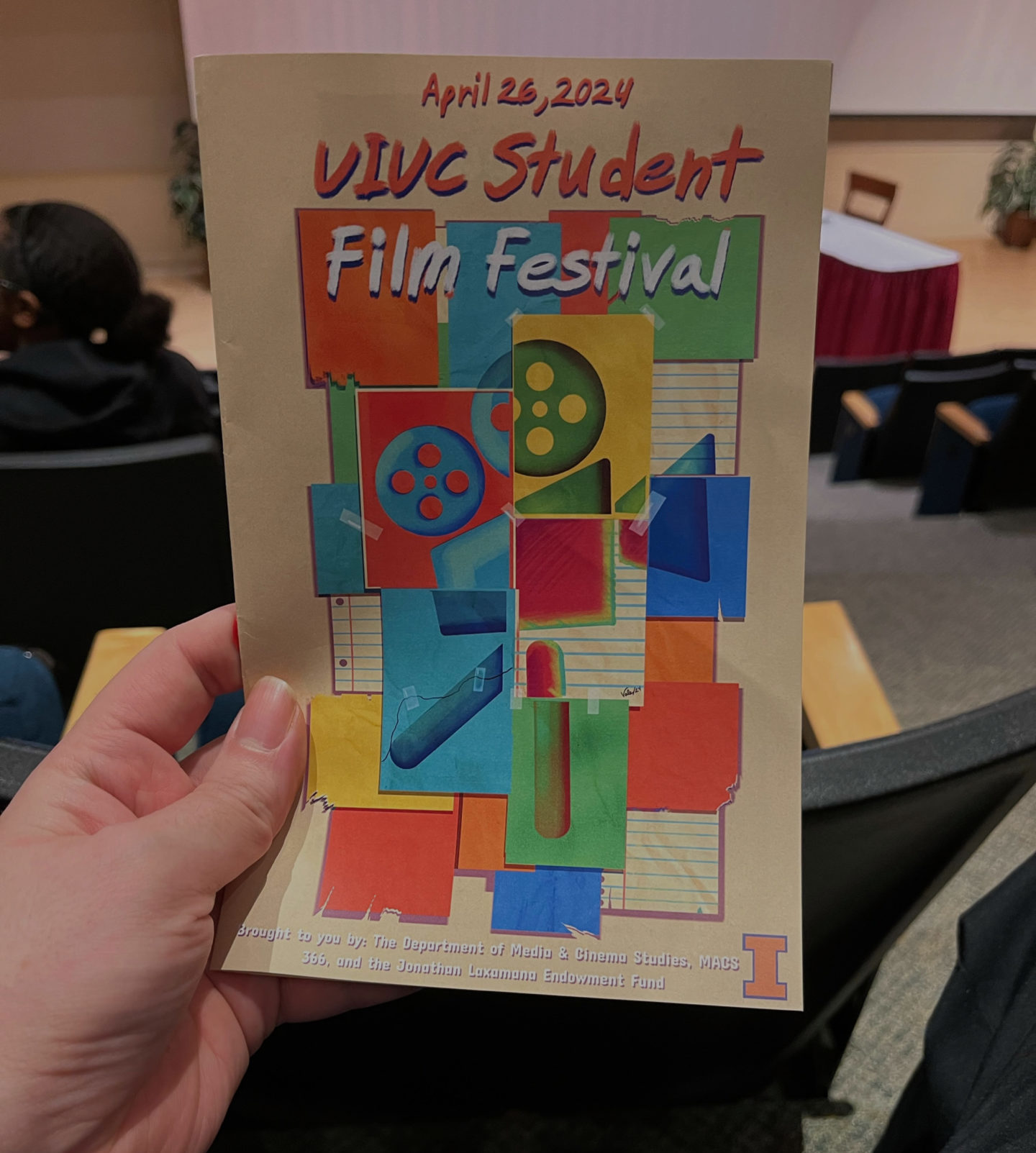 A white hand is holding a UIUC Student Film Festival program. The program has blocks of blue, yellow, and red with abstract images.