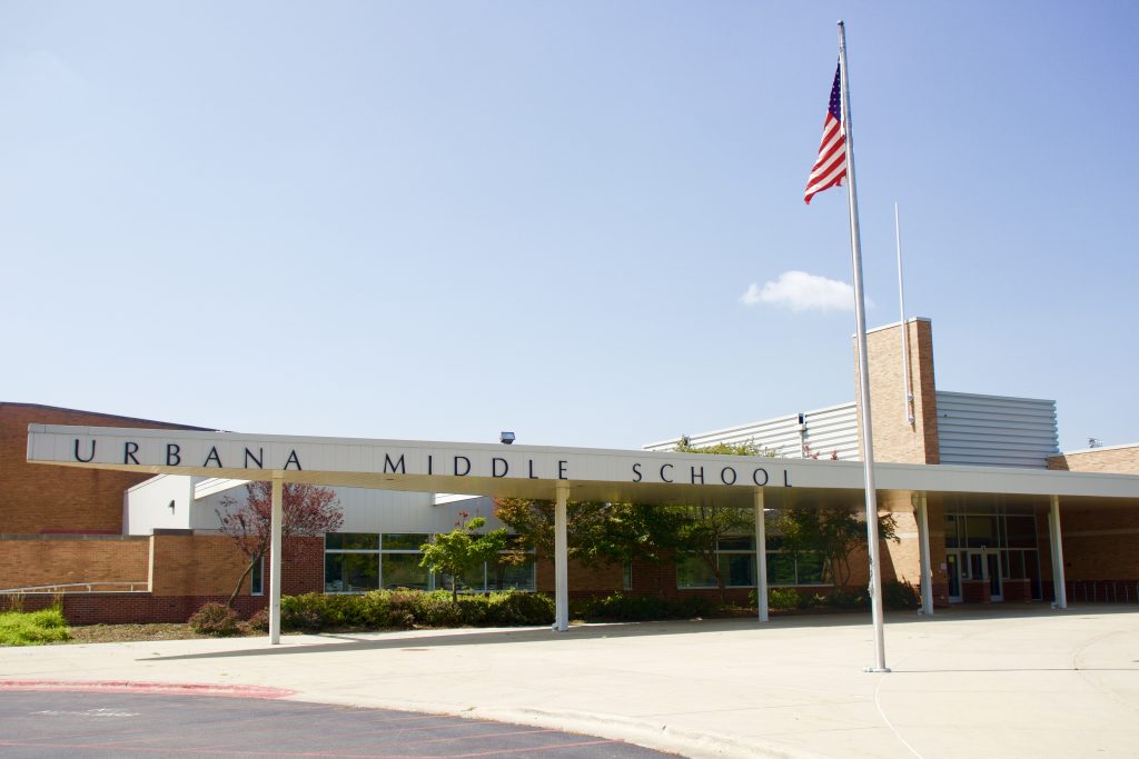 Photo of Urbana Middle School entrance. Background is a blue sky, there is an American flag to the front, right of the image. The building is brick with a white awning with the school's name written on the awning in brown, capital letters.