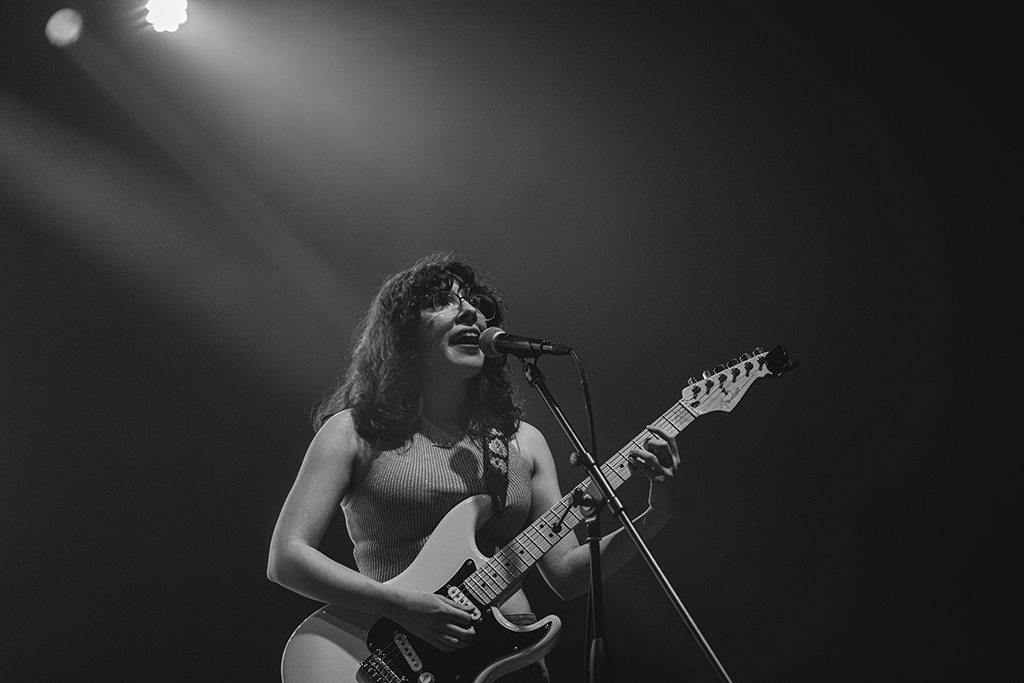 A musician with curly hair plays a white electric bass guitar and sings into a microphone on stage. The stage lighting casts a moody ambiance, suggesting a soulful and engaging musical performance.