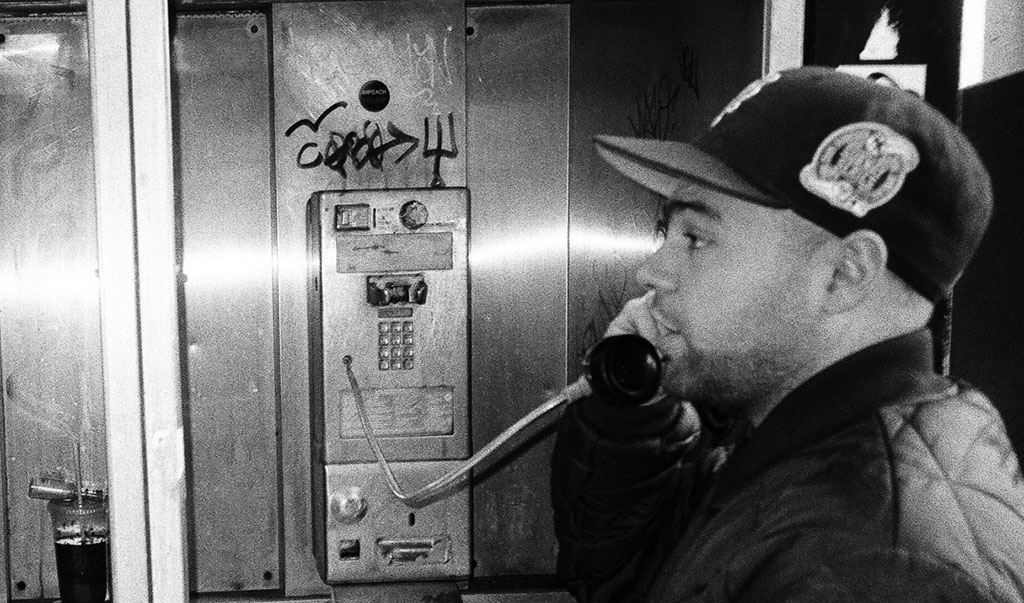 A grayscale snapshot of an individual using a public payphone. The scene has a gritty, urban feel, marked by graffiti on the phone booth. The person is wearing a baseball cap and jacket, indicative of a casual, street-style fashion. They are captured mid-conversation, which adds a candid and potentially urgent narrative to the photo.