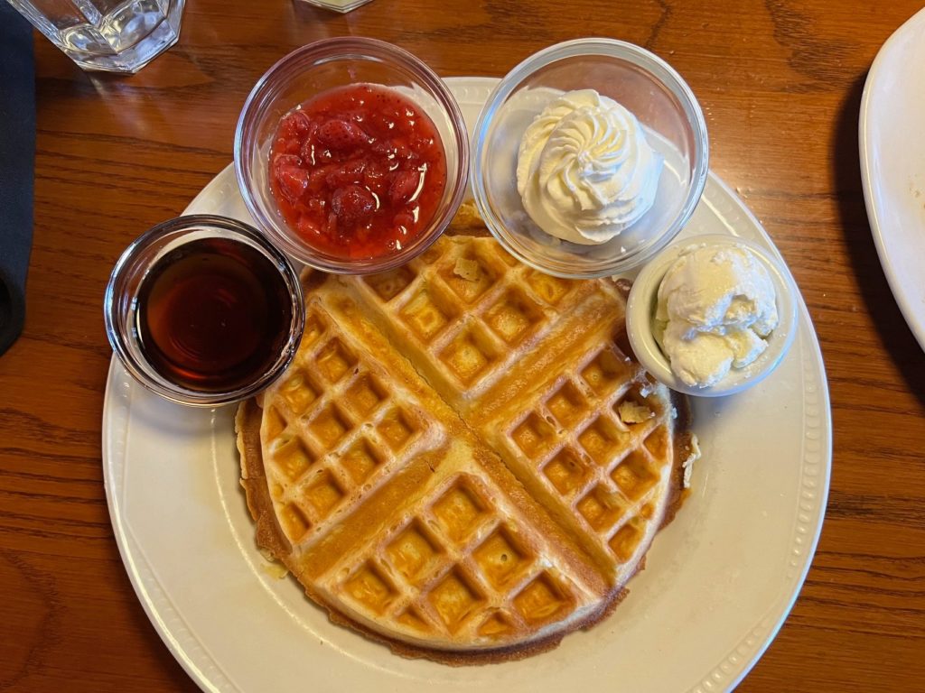 A waffle with side cups of syrup, strawberries, whipped cream, and butter.