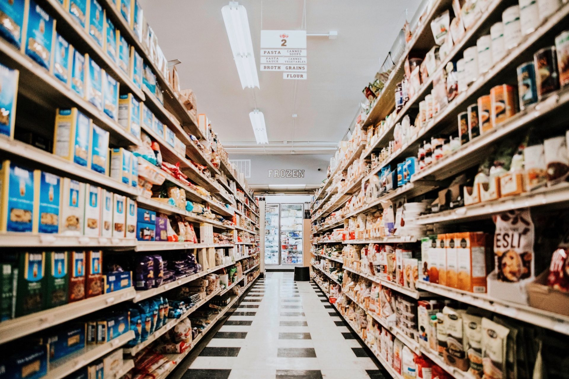 Aisle in a grocery store with products on shelves on both sides, a black and white checkered tile