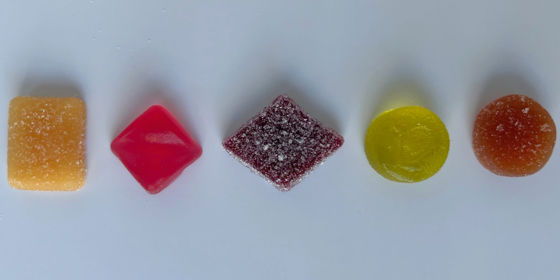 Five cannabis gummies in a row: orange square, pink diamond, purple diamond, yellow-green circle, and a redish pink circle.