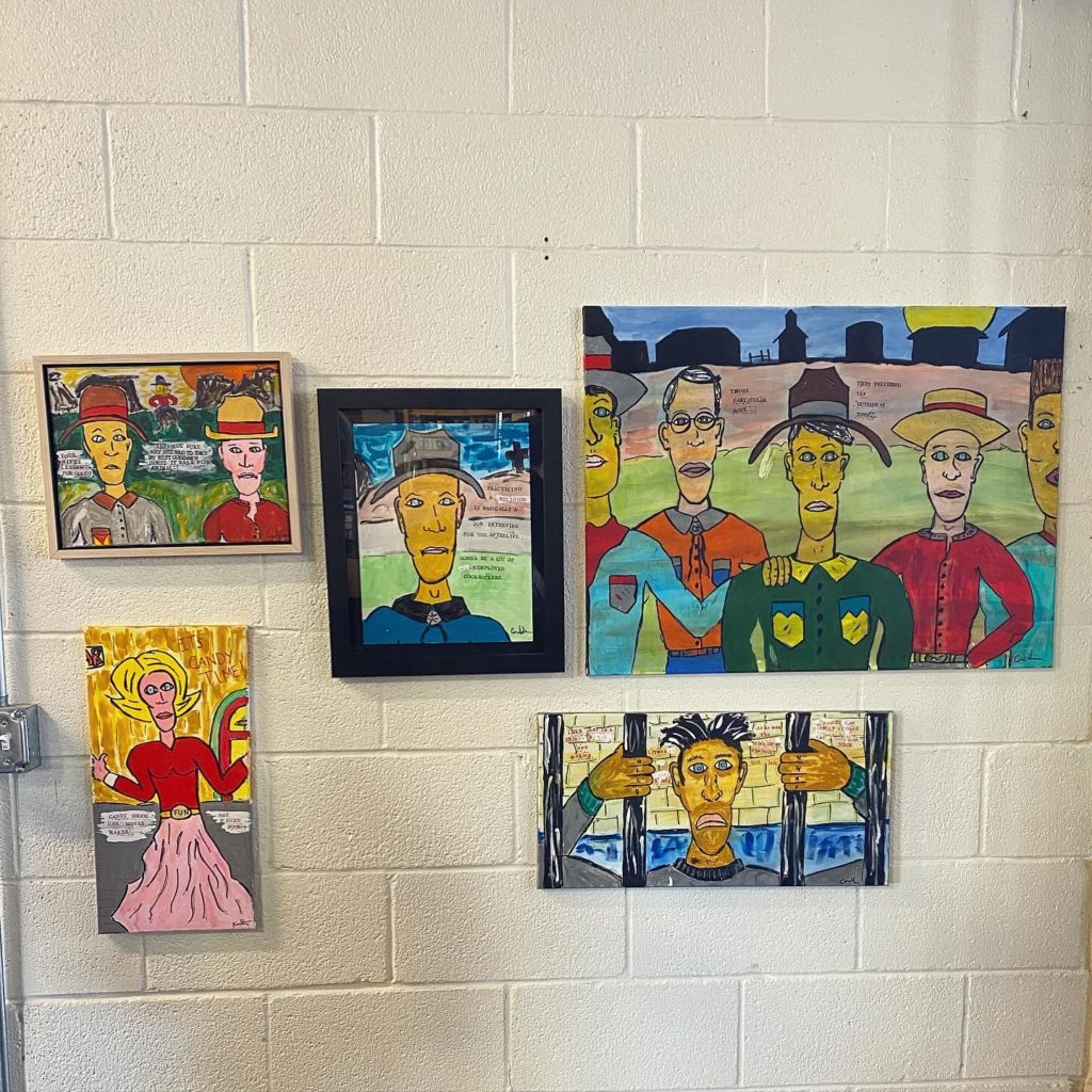 Five canvas prints hang on a white wall. The prints are colorful comic-book drawings of figures in jail, on a farm, and in front of a bright yellow window.