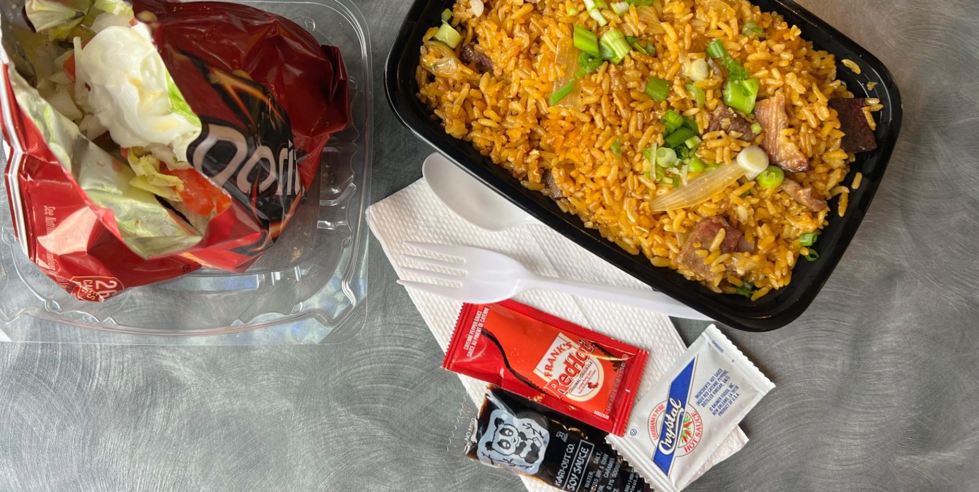 A Doritos bag has sour cream and taco ingredients beside a black container of fried rice topped with green onions. Beside both is a white paper napkin, white plastic fork and spoon, Frank's hot sauce, Crystal hot sauce, and soy sauce packet.