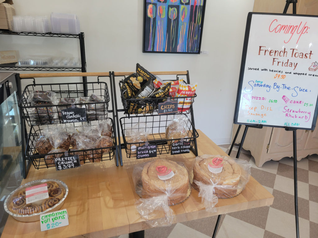 A counter holding cinnamon rolls, cakes, vegan brownies, pretzel crunch, chips and breadcrumbs with a sign nearby showing what food is coming up.