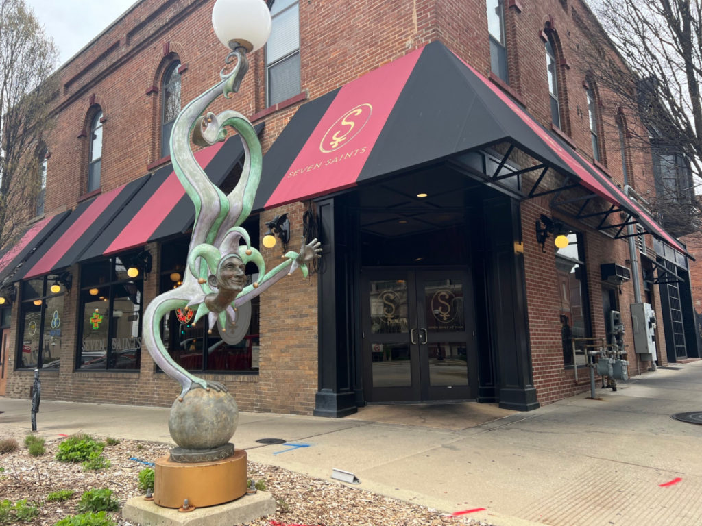 The exterior of Seven Saints on a cloudy day in Downtown Champaign. The lightpost is a statue of a jester standing on one hand.