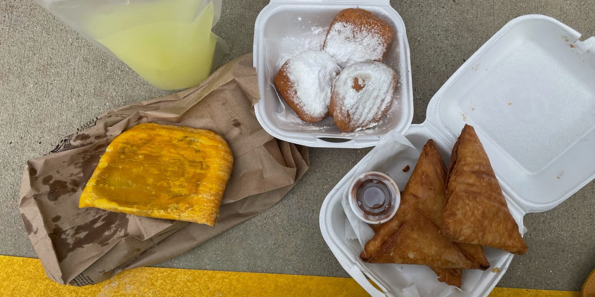 The author's order at Stango Cuisine: ginger juice in a plastic bag, a beef handpie on a brown paper bag, a white styrofoam container of beignets, and three somaosas with a side sauce.