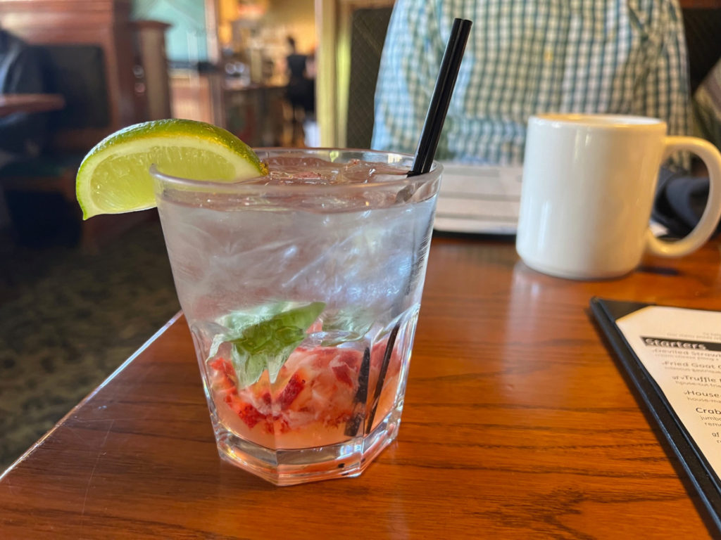 A glass cup with clear liquid and basil and strawberries on the bottom, garnished with a lime wedge.