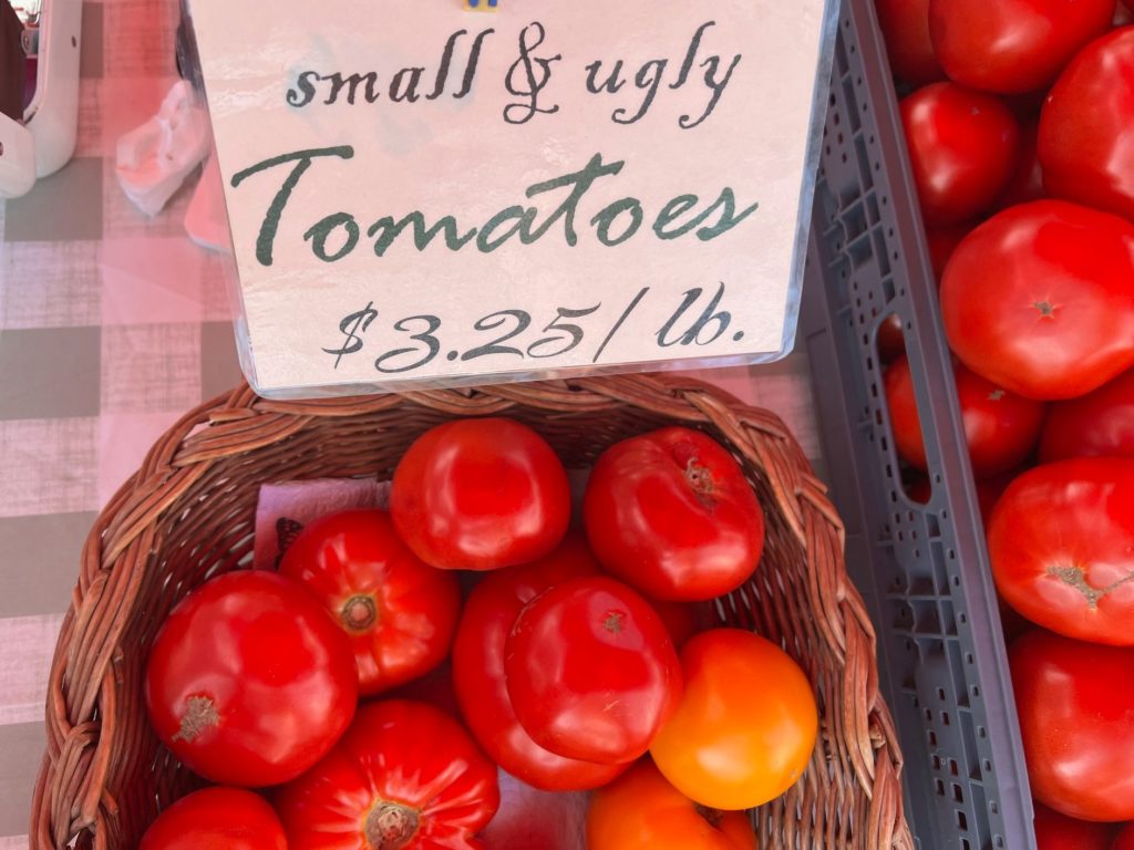 Many oddly shaped tomatoes for sale in a brown basket.