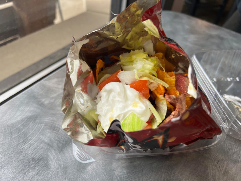 A walking taco with sour cream, chopped iceberg lettuce, tomato, brisket, and red Doritos chips inside a Doritos snack-sized bag inside of a clear clamshell takeout container on a silver table by a window.