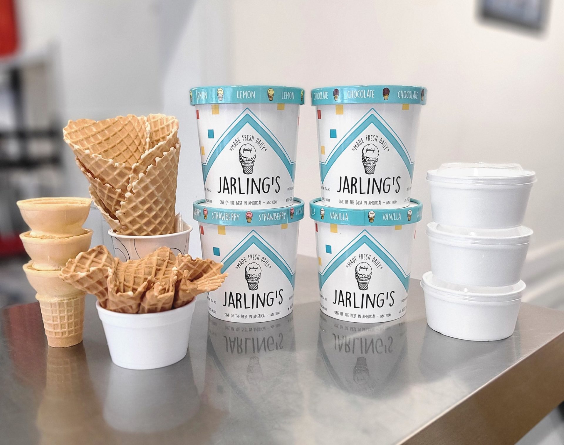 Four quarts of ice cream next to two waffle cones and white styrofoam cups
