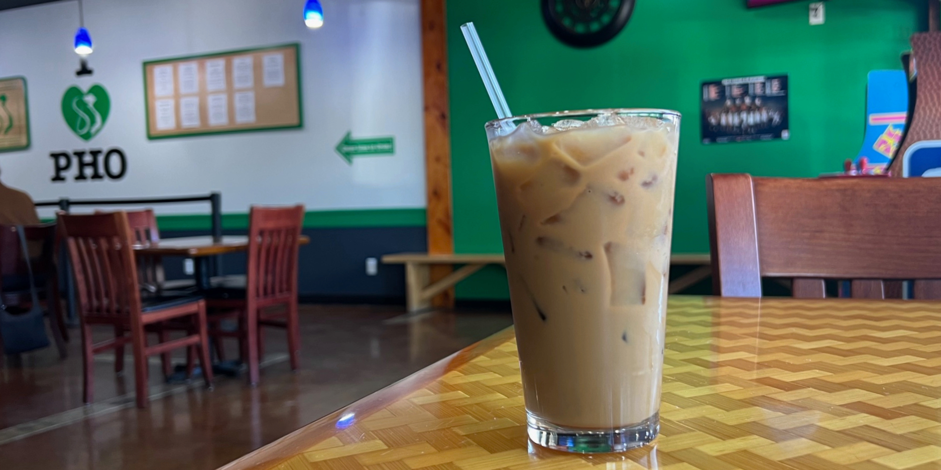 A pint glass has a light brown liquid with big flat square ice cubes and a clear straw. The dining room has a white wall that says "I heart pho" and there is a bright green accent wall to the right.