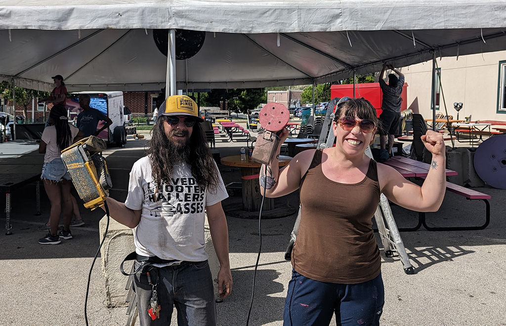 Two individuals in a celebratory mood, likely involved in setting up or participating in the event under the tent. A man with long hair, a beard, and a cap, is holding a yellow sander, while a woman, sporting sunglasses, poses energetically with sanding equipment. Their attire and equipment suggest they might be part of the crew preparing the venue.