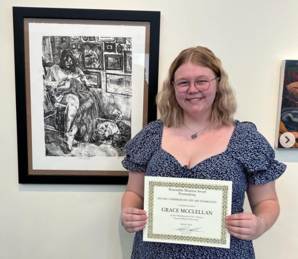Photo of a white woman with blonde hair and glasses stands in front of a black and white lithograph artwork.