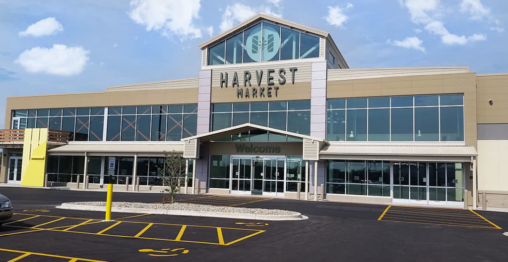 The exterior or the Harvest Market grocery store. It is two story stone and glass windows with a pointed roof in the middle of the building. There is a parking lot just outside the double doors.