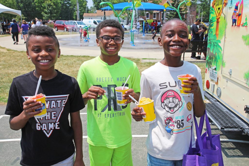 Three Black boys smile at the camera holding snow cones, a splash pad is in the background.