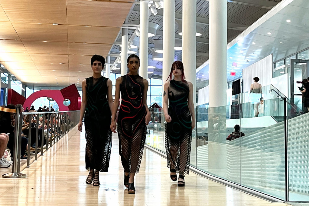 Three woman walk the runway at ReFashioned. They are all wearing different versions of a black dress with mesh skirts. The woman in the middle wears a dress with red details.