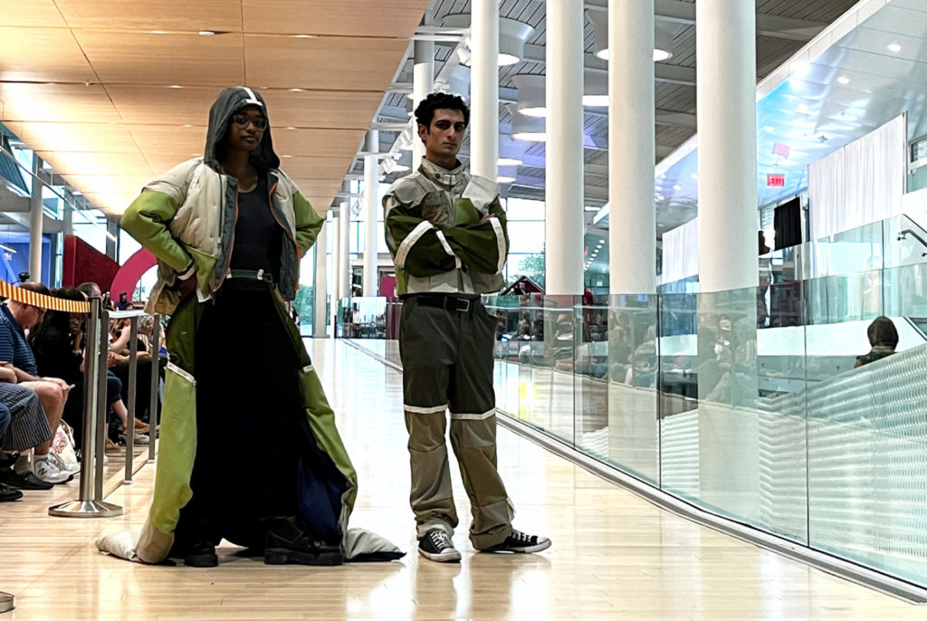 Two models, one Black woman in glasses on the left, and a Brown man on the right. They wear monochromatic outfits in different shades of green. The woman wears a hood and long cape / train, and the man wears pants and a coat.